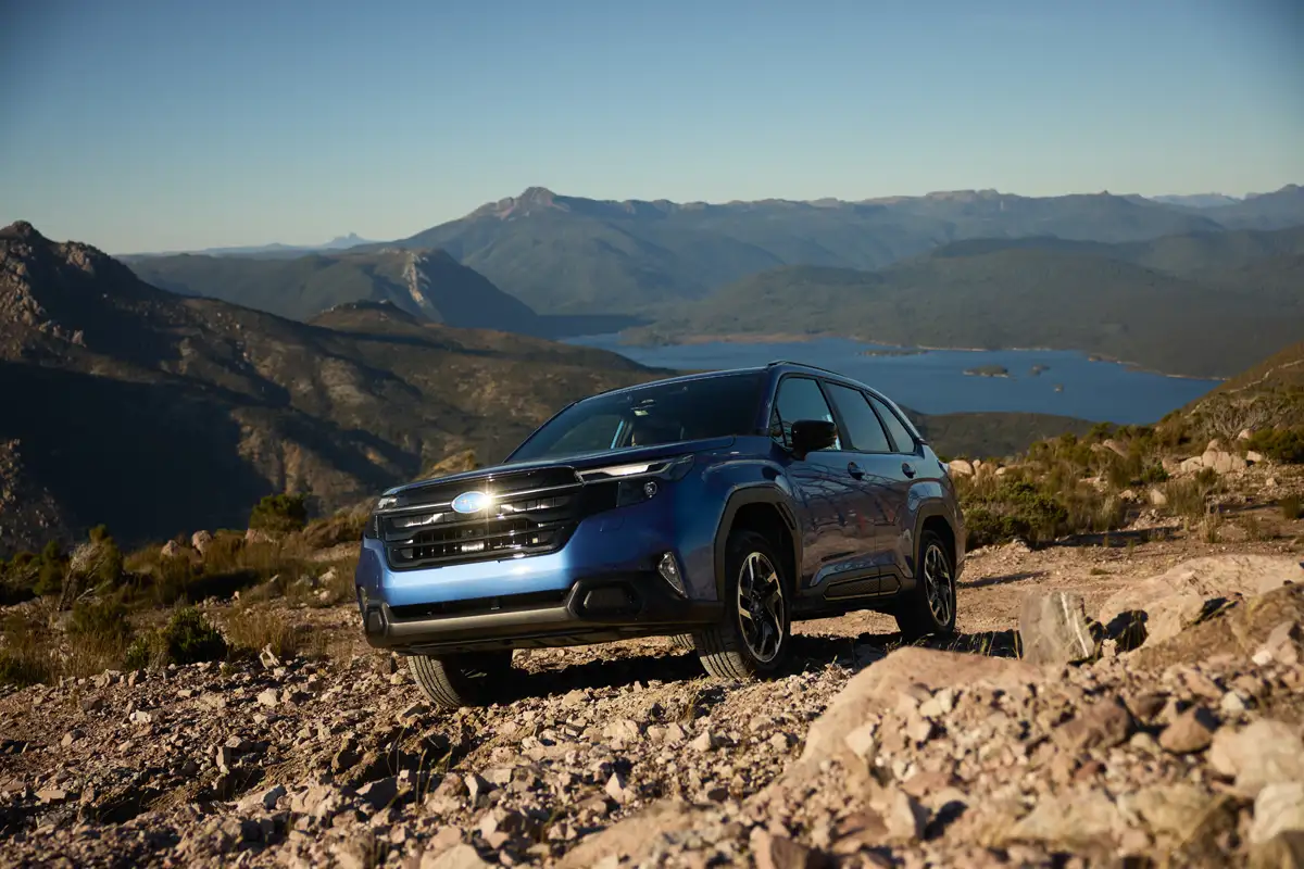 Xe ô tô SUV Subaru Forester màu xanh chạy trên địa hình đồi núi đá sỏi gồ ghề phía trước hồ nước lớn.